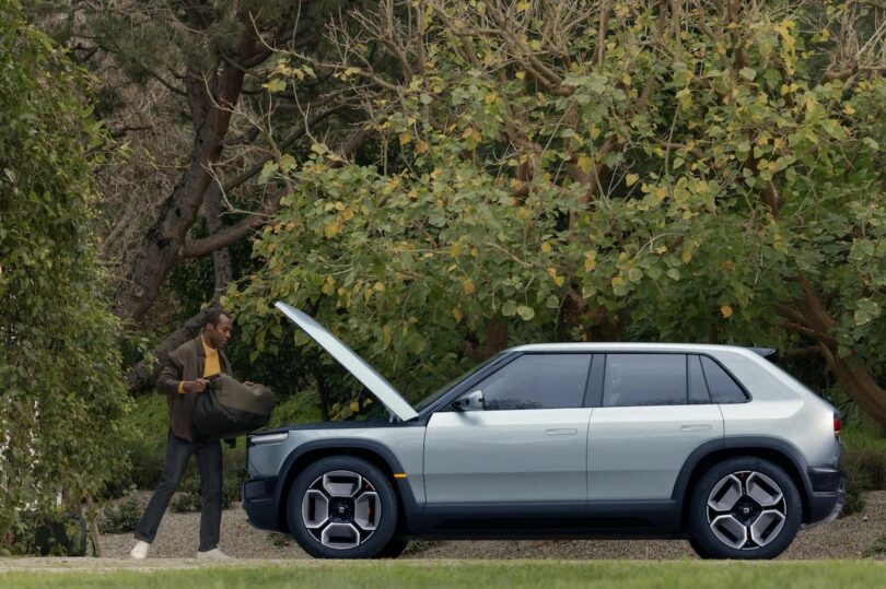 A man loading luggage into the trunk of a modern silver suv parked by lush greenery.