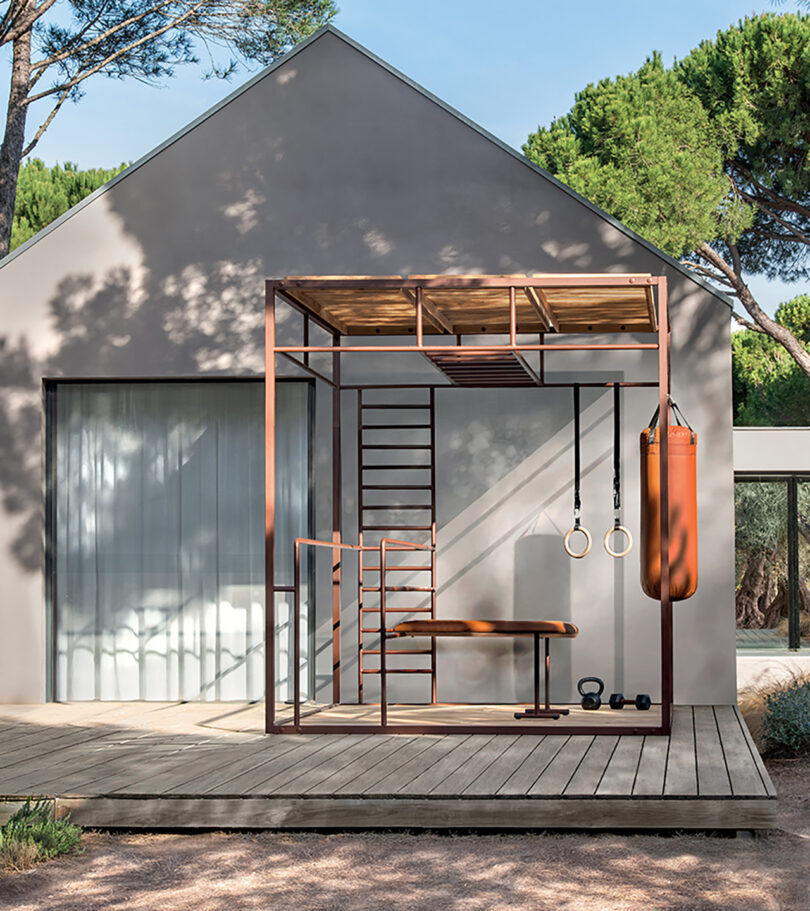 Modern house with an OUT-FIT outdoor gym area set on a wood deck, equipped with a punching bag, gymnastic rings, and weights on a wooden platform surrounded by trees.