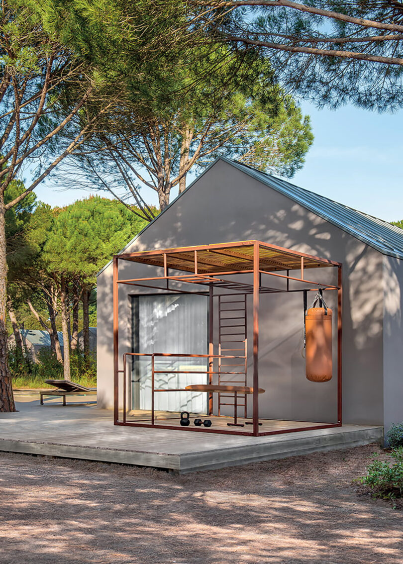 Modern metal frame and teak slat OUT-FIT outdoor gym set in front of a minimalist gray residence with large glass doors, surrounded by tall green trees.