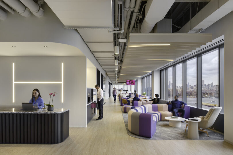 Modern office lounge area with people relaxing and working, featuring stylish furniture, large windows with a city view, and a reception desk