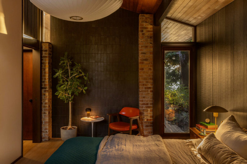 Cozy bedroom featuring a bed with blue and white bedding, a wooden floor, dark textured walls, and a large window. there is a round white pendant light and plants in the room.