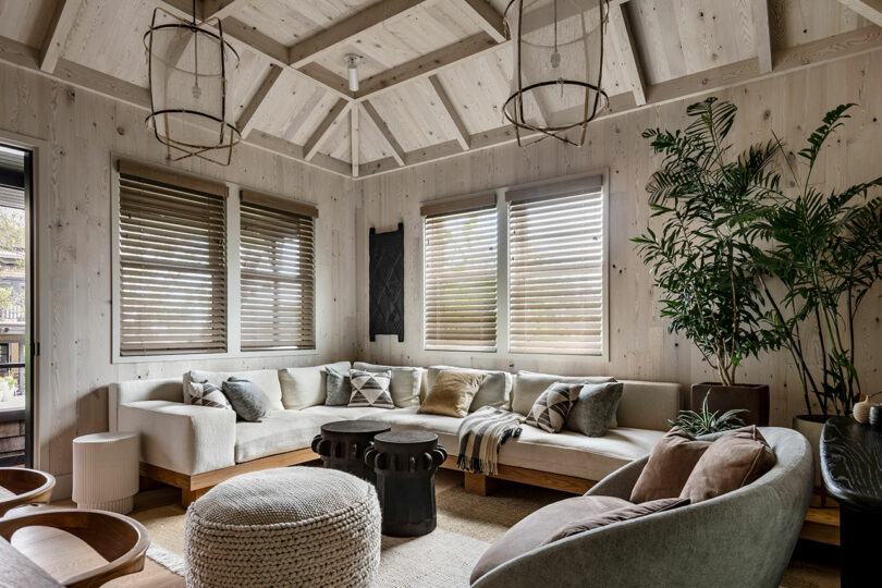 Modern living room designed by SkB Architects, featuring a beamed ceiling, large-cushioned sectional sofa, pendant lights, and lush indoor plants near shaded windows.