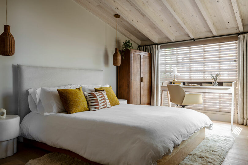 Cozy bedroom designed by SkB Architects with a white bed, wooden ceiling, hanging wicker lamps, and a desk by the window, conveying a calm, natural aesthetic.