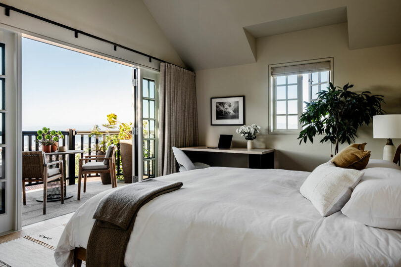 A stylish bedroom designed by SkB Architects with white bedding, featuring a balcony with outdoor seating, large windows, and tasteful decor.