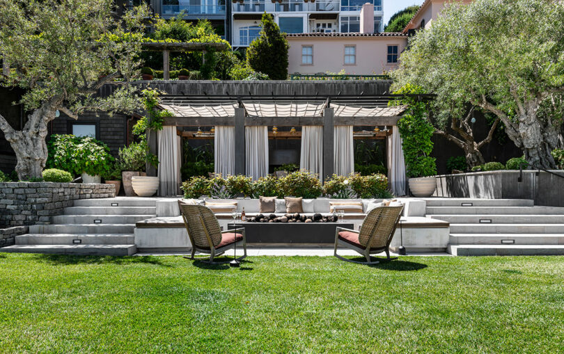 Outdoor seating area designed by SkB Architects with two wicker chairs facing a cushioned bench under a pergola, surrounded by lush greenery and stone steps, with a building in the background.