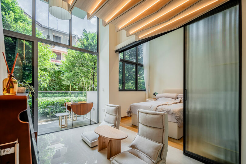 Modern bedroom with large windows, a bed, two chairs, and a desk, overlooking a garden with lush greenery.